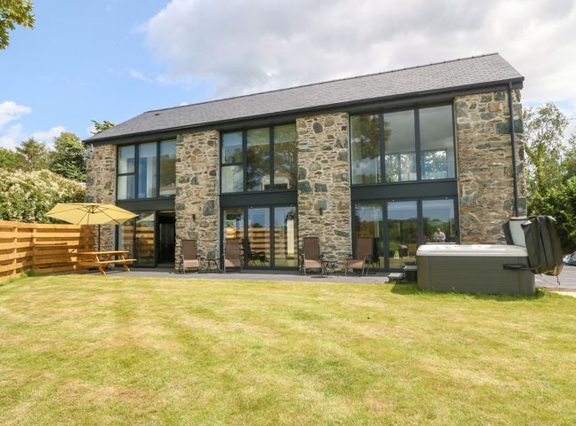 A stone house with large windows and doors a grassy yard with patio chairs a wooden fence and a hot tub at Pen-Y-Bryn in Llanfairpwllgwyngyll