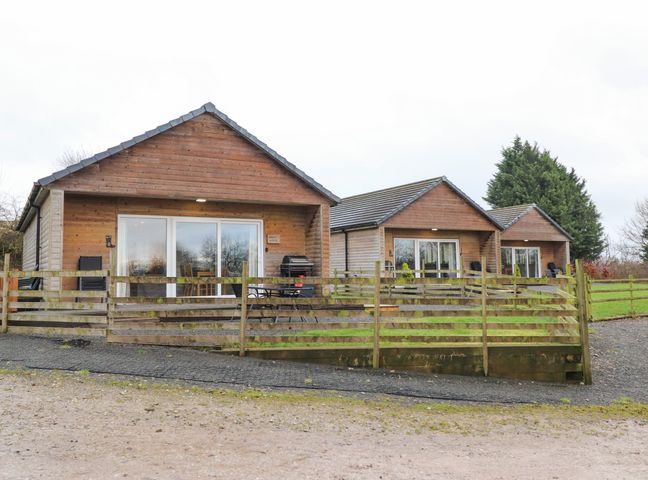 Three wooden lodges with glass sliding doors and fenced patios on a gravel surface at Holly Lodge in Goosnargh