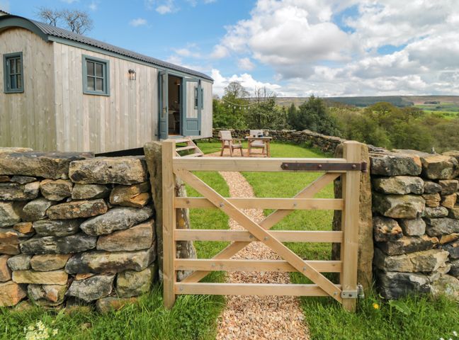 A wooden shepherds hut with an open door behind a wooden gate and stone wall with chairs and table on grass at Sheep Cote Shepherds Hut in Chop Gate near Stokesley
