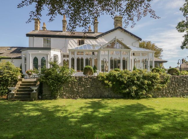 A backyard with grass bushes and stone wall in front of a white house with multiple chimneys and a glass conservatory at Valley View in Dalton-In-Furness