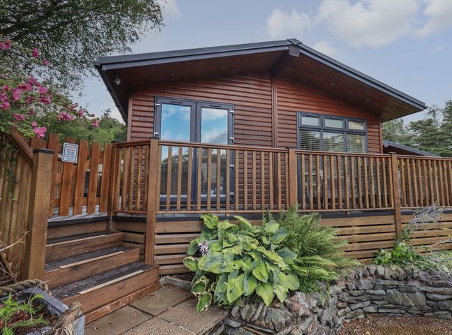 A wooden cabin with a deck and railing surrounded by plants at Hill Top Lodge in Bowness 65 White Cross Bay