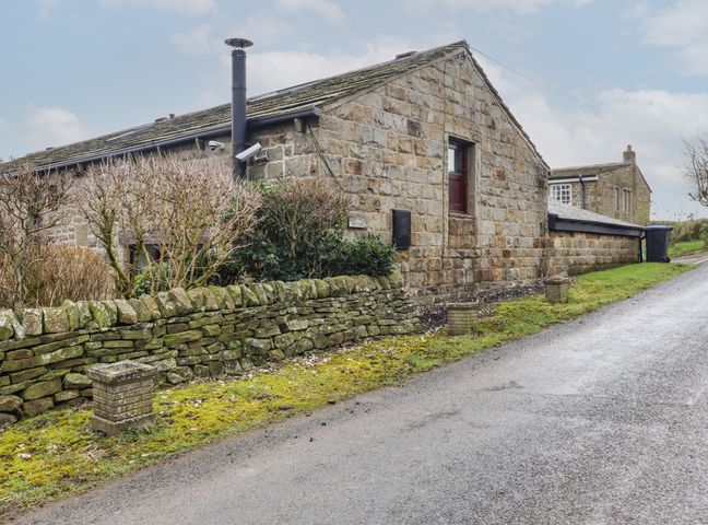 A stone building with a mossy stone wall and trimmed trees beside a road at Bride Cross Granary in Otley