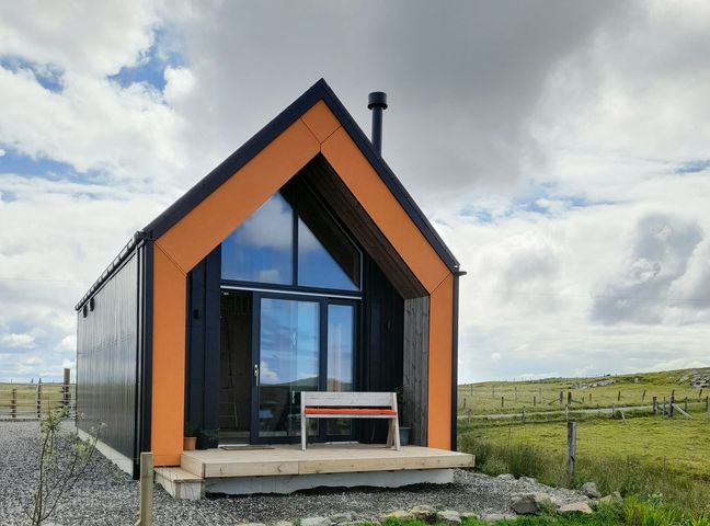 A small modern cabin with orange and black accents and a bench on the porch in a grassy rural area at North Light Studio in Locheport near Lochmaddy North Uist