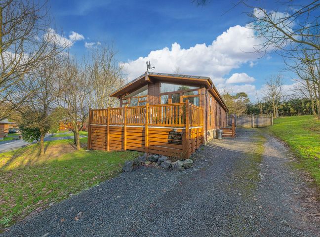 A wooden cabin with a fenced deck and gravel driveway surrounded by trees at Fern Lodge in Allithwaite near Cartmel