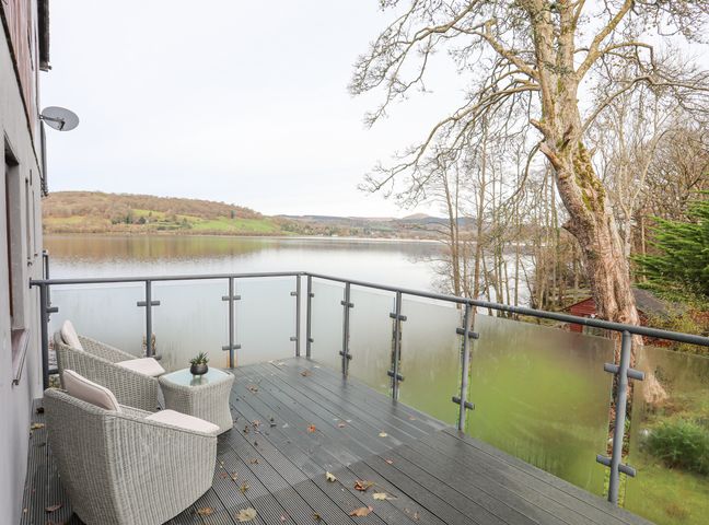 A balcony with two chairs a small table and glass railing overlooking a lake and hills at Vyrnwy Lakeside Bala Lake