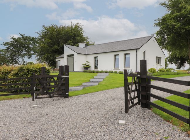 A white single-story house with a gray roof behind a wooden gate on a gravel driveway at Shannon Vale in Woodford County Galway