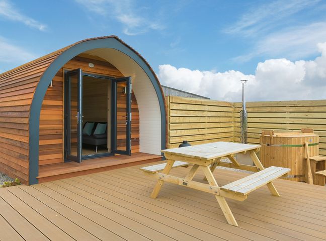A wooden pod with open doors next to a picnic table and a wooden hot tub on a deck at Sun Rise in St Buryan