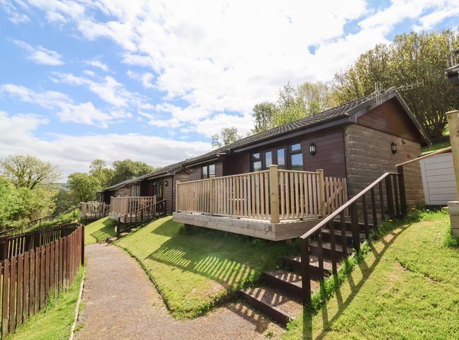 A row of single-story chalets with wooden railings along a pathway surrounded by grass at Chalet Log Cabin L14 in Combe Martin