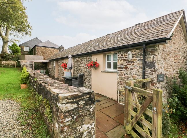 An exterior view of a stone cottage with a wooden gate and outdoor seating at Cosy Cottage in Broadwoodwidger near Lifton