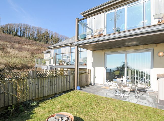 A garden patio with a glass table and chairs outside a house with glass balcony at Pebbles in Downderry