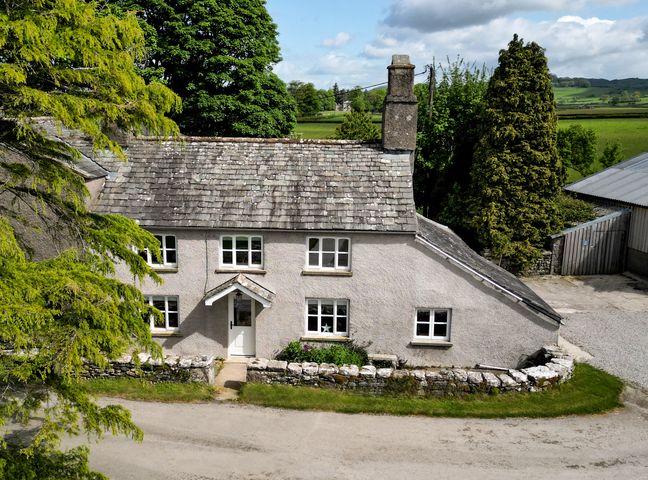 A stone house with a tiled roof surrounded by trees and greenery at Longlands Farm Cottage in Cartmel