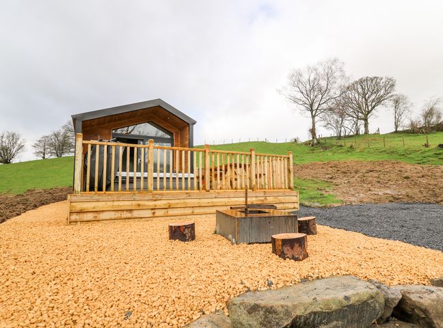 A small wooden cabin with a patio and hot tub on a gravel area with tree stump seats and green grass hill in the background at Fawn's Rest in Nantmel near Rhayader