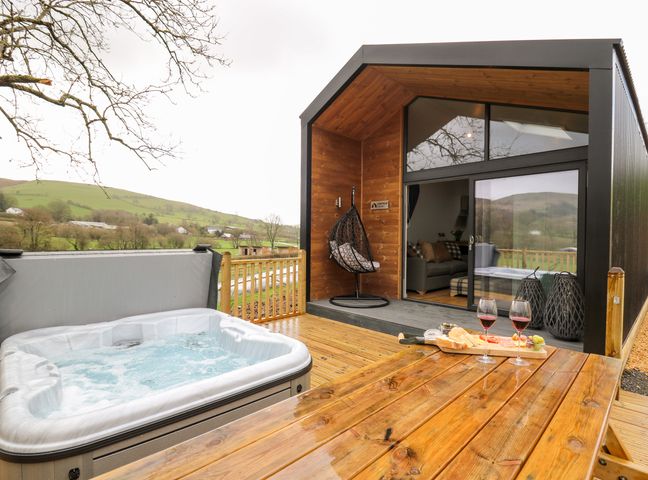 A wooden cabin with a hot tub and a picnic table with wine glasses and snacks on the deck at Doe Lodge in Nantmel near Rhayader