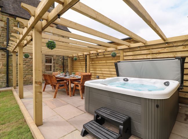 An outdoor patio with a hot tub a dining table with chairs and hanging plants at Upper Hurst Farmhouse in Hulme End near Hartington