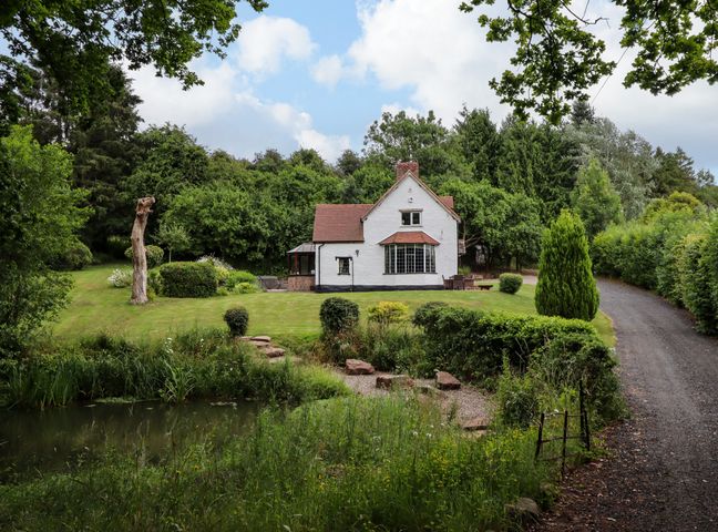 A white house with a red roof surrounded by greenery and a pond in front at Grove Cottage in Leominster