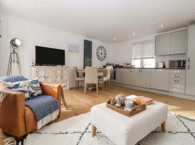 An open plan living room and kitchen with a leather armchair a white ottoman a round dining table and grey kitchen cabinets at The Gallivant in Southwold