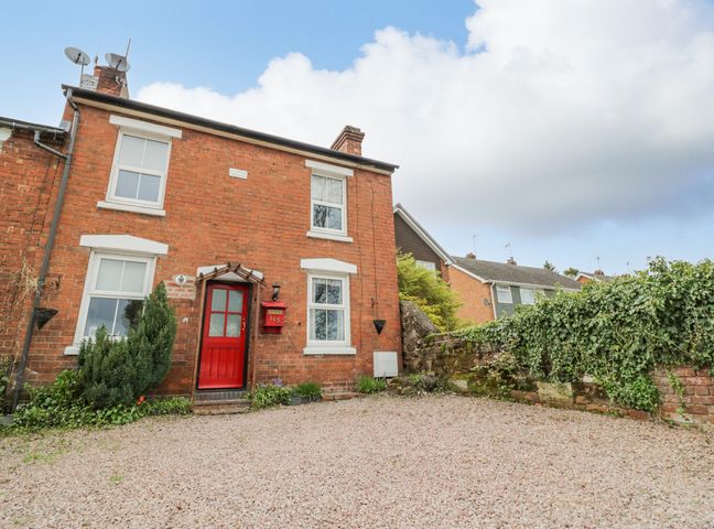 A brick house with a red door and mailbox surrounded by a gravel driveway and greenery at Dobbies Den in Bewdley