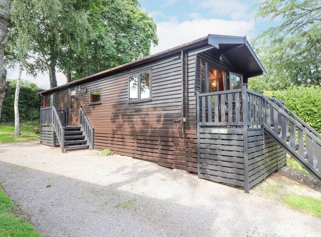 An exterior view of a brown wooden lodge with steps and railings surrounded by trees and grass at Ennerdale Lodge in Burnside Park near Keswick