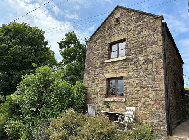 A stone house with two windows and two wooden chairs outside surrounded by green bushes and trees at Old Post Office Barn Upper Hulme near Leek