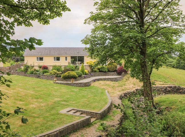 A garden with a lawn large tree stone walls and shrubbery with a yellow building in the background at Llety'r Bugail in Glynneath