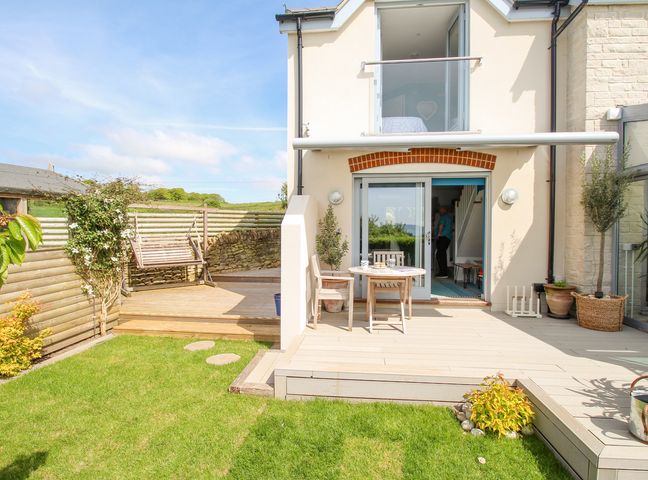 A garden with grass a wooden deck with a swing and a patio with chairs and a table at Blue Waters in Weymouth