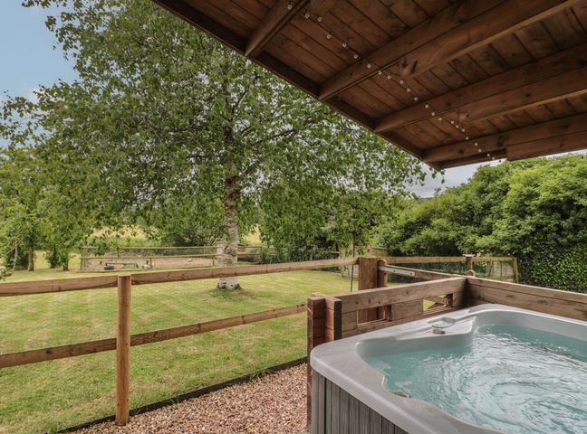 An outdoor hot tub under a wooden roof with a grassy yard and trees at Pigsfoot Lodge in Ash Thomas near Halberton Devon