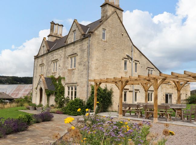 A stone farmhouse with a wooden pergola and outdoor seating surrounded by flowers and greenery at Hammonds Farmhouse in Stroud