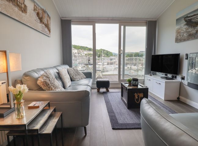 A living room with a gray sofa, coffee table, television, lamp, and a view of boats outside at Moelwyn  57D South Snowdon Wharf in Porthmadog