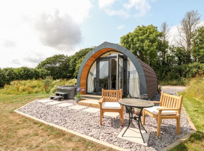 A small cabin with wooden furniture and a table outside on a gravel patio surrounded by grass and trees at Oak in Elmgate near Saltash