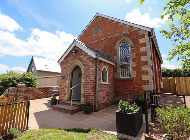 A brick building with arched windows and a small porch with steps surrounded by plants at The Old Chapel in Dorstone near Hay-on-Wye