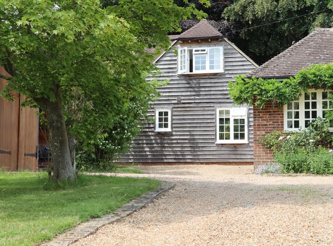A gravel driveway leading to a house with wooden and brick exterior surrounded by trees and plants at Spindlewood Cottage in Hawkhurst
