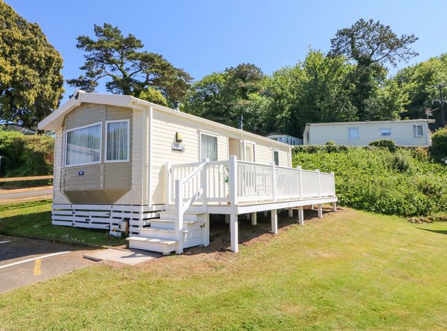 A beige mobile home with a white railing on a grassy slope with trees behind at Horizon in Combe Martin
