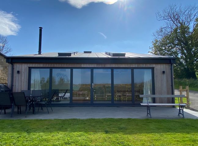 A wooden building with large glass doors a patio table with chairs and a bench on a lawn at 1 Orchard Cottages Dunstan near Craster