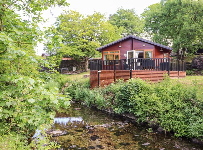 A wooden lodge beside a clear stream surrounded by green trees and shrubs at Nutkins Lodge in Beckside 5 Limefitt Park near Windermere
