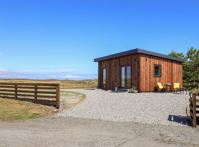 A small wooden cabin with a gravel driveway and two yellow chairs outside at The Wheelhouse in Grimsay near Balivanich