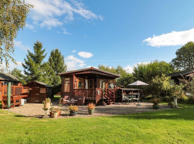 A wooden cabin with a deck surrounded by potted plants lawn and outdoor seating at Evergreen Lodge Felmoor Holiday Park near Felton