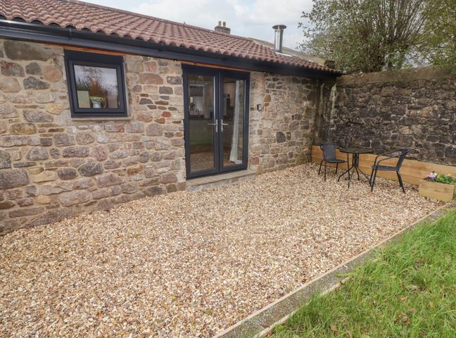 A stone building with tiled roof and glass door opening to a gravel patio with a small table and two chairs at The Nest in Amroth