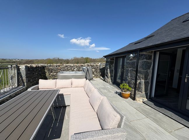 An outdoor patio with a sectional sofa and table next to a stone building and a stone wall with a hot tub behind Gaerau Bach in Llanaber