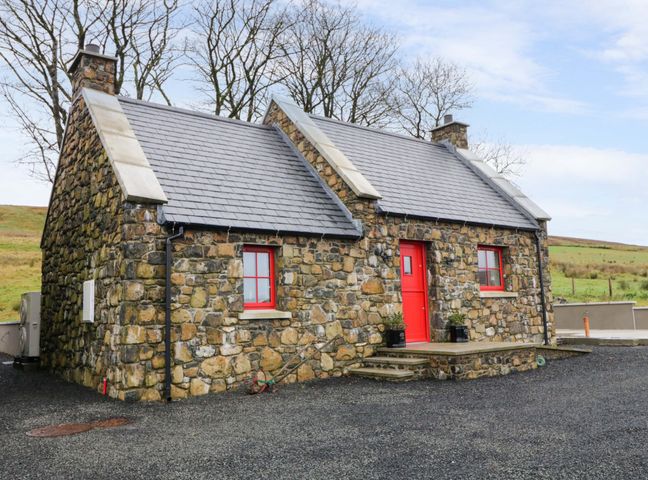 A stone cottage with a red door and windows surrounded by a gravel driveway and trees at The Croft in Newtown-Crommelin near Cloughmills