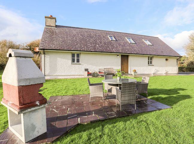 An outdoor patio with a table and chairs next to a barbecue grill in front of a house with skylights and a wooden door at Rhyd Y Bont Bach in Rhoscolyn