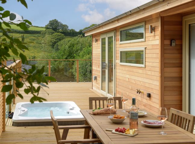 A wooden outdoor deck with a hot tub and a dining table set with food and drinks at 4 Valley View in Lanreath