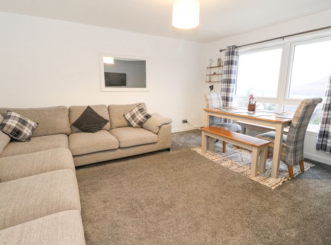 A living room with a beige sectional sofa and a wooden dining table with plaid chairs and bench at 22 Moray Place in Fort William
