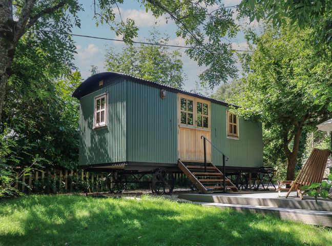 A green shepherd hut on wheels with wooden doors and steps in a garden with trees and lawn at The Happy Hut in Sopley near Christchurch