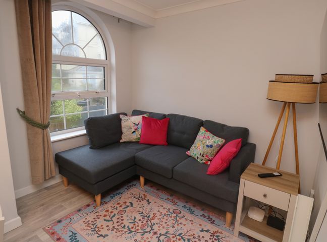 A corner sofa with cushions beside an arched window with curtains a wooden floor lamp and a side table with a remote on a rug at 1 Ilsham Cottages in Torquay