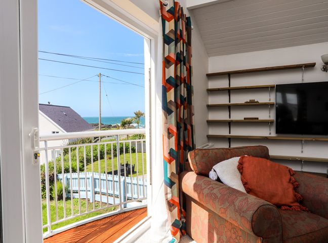 A room with a patterned armchair and cushions next to a glass door leading to a balcony overlooking a garden and the sea at Seaside Sanctuary in Trearddur Bay