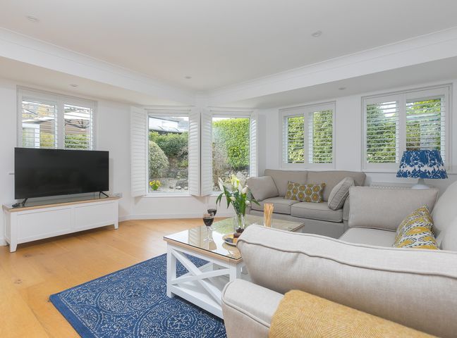 A living room with two beige sofas a coffee table a television and windows with white shutters at Lowena in Carbis Bay