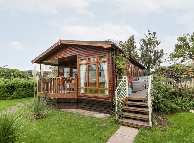 A wooden cabin with a porch and stairs surrounded by green grass and plants at Eden Lodge in Castlewigg near Whithorn