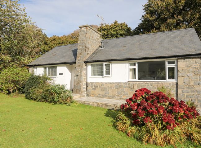 A stone and white exterior cottage with a large chimney and garden with flowers and shrubs at Ty Cerrig in Llanbedrog