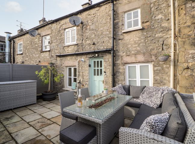 An outdoor patio with a wicker sofa and table set beside a stone building with windows and a mint green door at Market Square Maisonette in Kirkby Lonsdale
