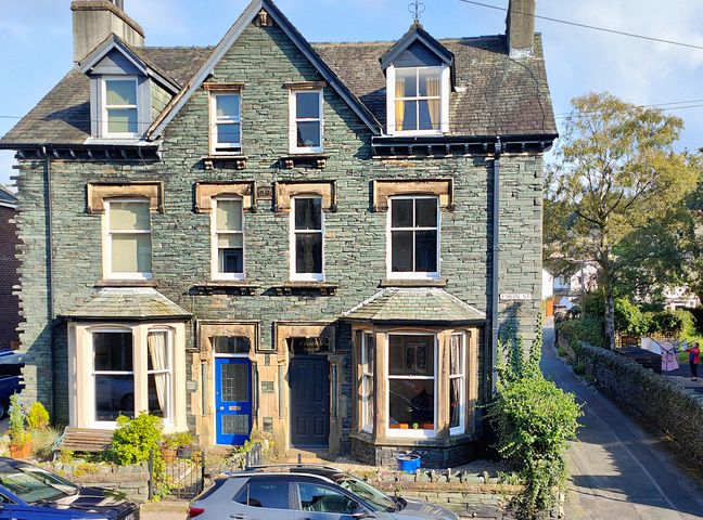 A stone house with two front doors and multiple windows on a street with parked cars at Cranford House in Keswick